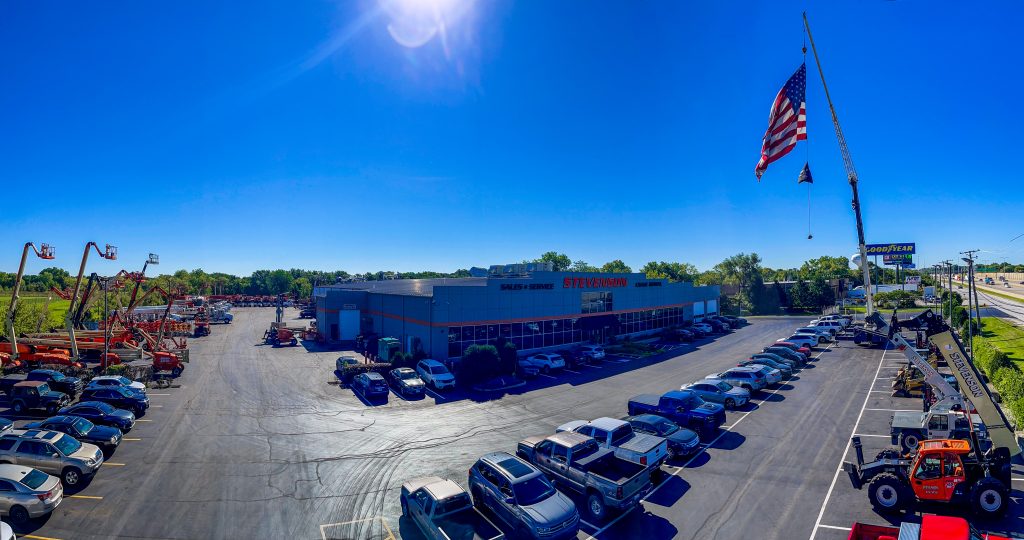 A panoramic view of Stevenson Crane’s headquarters with rows of heavy equipment, customer parking, a large crane hoisting an American flag, and clear blue skies above.